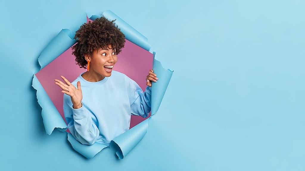 Overjoyed African American woman raises palm and looks gladfully aside as meets someone wears casual jumper poses through paper background in hole expresses positive emotions.